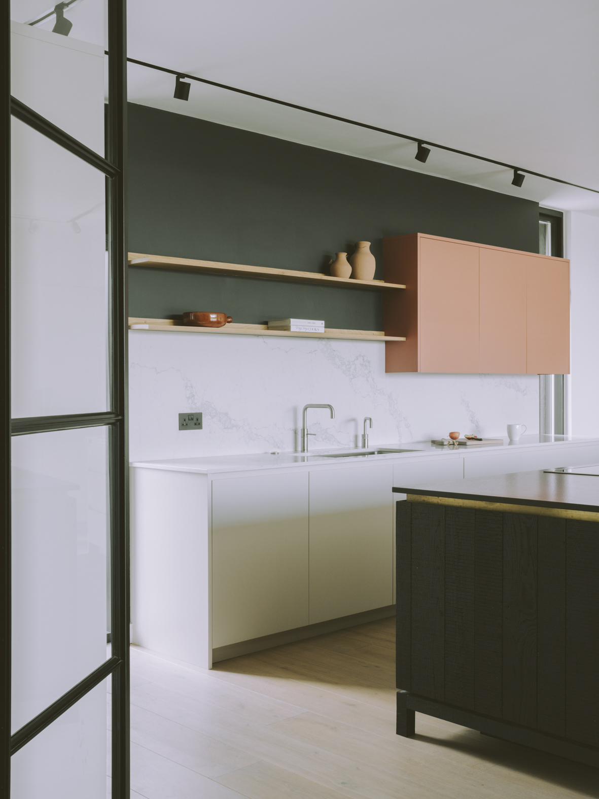 Chelwood house's kitchen with a black island, surrounding white units, pink wall units and open shelving.